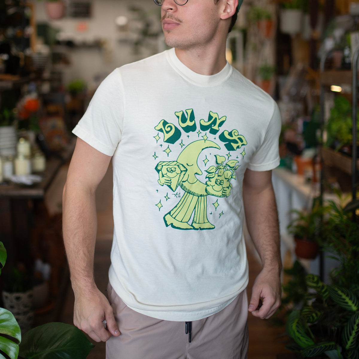 A man wearing a white graphic t-shirt with a green cartoon moon and sun design, paired with a green baseball cap and glasses, standing in a plant-filled indoor shop.